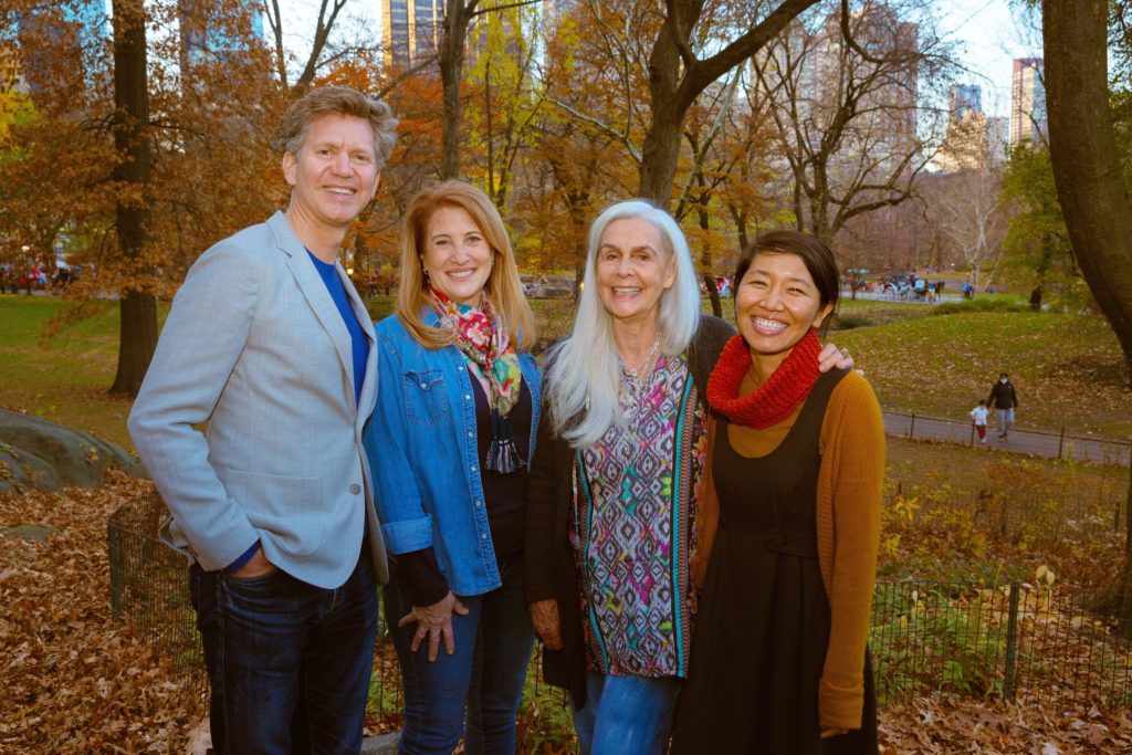 A man and three women, smiling and standing in a city park with trees and autumn leaves