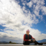 Dzongsar Khyentse Rinpoche sits on the ground wearing a pink hat with a big blue sky behind him.