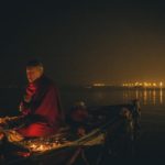 Dzongsar Khyenste Rinpoche is on a boat performing a fire puja in the nighttime, lights reflect off the water in the distance.