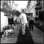 A gentle man stands in the center of a group of people with a spotted dog to his right.