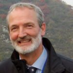 A grey haired bearded man smiles with a mountain landscape behind him.