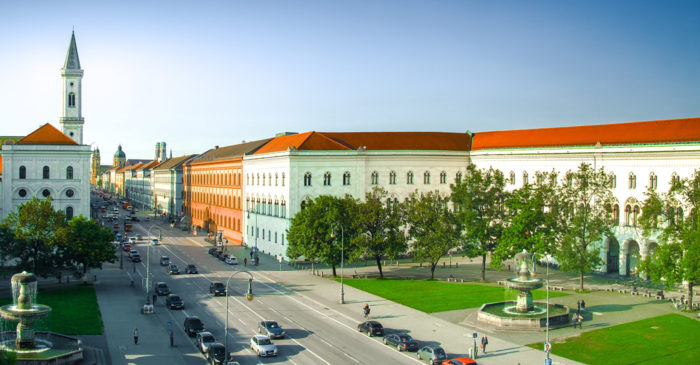 Old buildings with crimson roofs on both sides of a four-lane street. Fountains, green grass and trees on both sides.