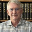 A white haired gentleman in a plaid shirt sits in front of a library of books.