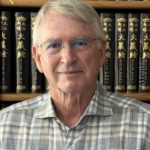 A white haired gentleman in a plaid shirt sits in front of a library of books.