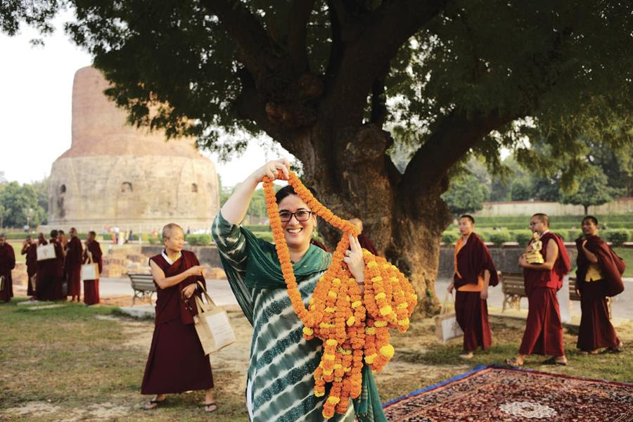 A woman smiling and holding marigold garlands. Red-robed monks behind her. The Sarnath stupa is in the background.