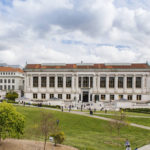 A huge, light-gray university building with a tall, clock-topped tower, a patch of green grass in front of it, and a blue sky in the distance. A large number of students are seen passing the building.