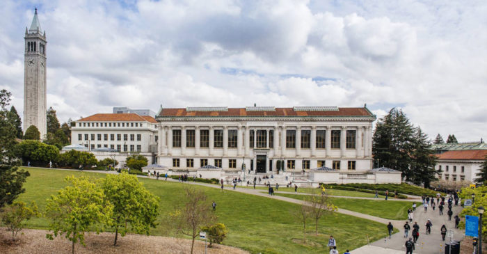 A huge, light-gray university building with a tall, clock-topped tower, a patch of green grass in front of it, and a blue sky in the distance. A large number of students are seen passing the building.
