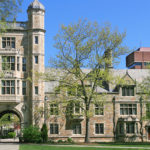 Large old brown university building with green trees in front of it and blue sky in the background.
