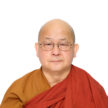 A monk wearing scarlet and gold colored robes wearing glasses sits in front of a white background.