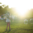 A girl is standing in a grassy field. The background is blurred, but shows some trees and buildings, and a bright sun, just behind the buidlings. The girl is facing toward the buildings, and appears to be turning. Her toes are pointed toward each other and she is holding a wand from which a dozen or so large head-sized bubbles have come, floating around her.