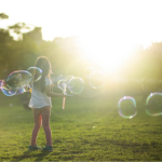 A girl is standing in a grassy field. The background is blurred, but shows some trees and buildings, and a bright sun, just behind the buidlings. The girl is facing toward the buildings, and appears to be turning. Her toes are pointed toward each other and she is holding a wand from which a dozen or so large head-sized bubbles have come, floating around her.