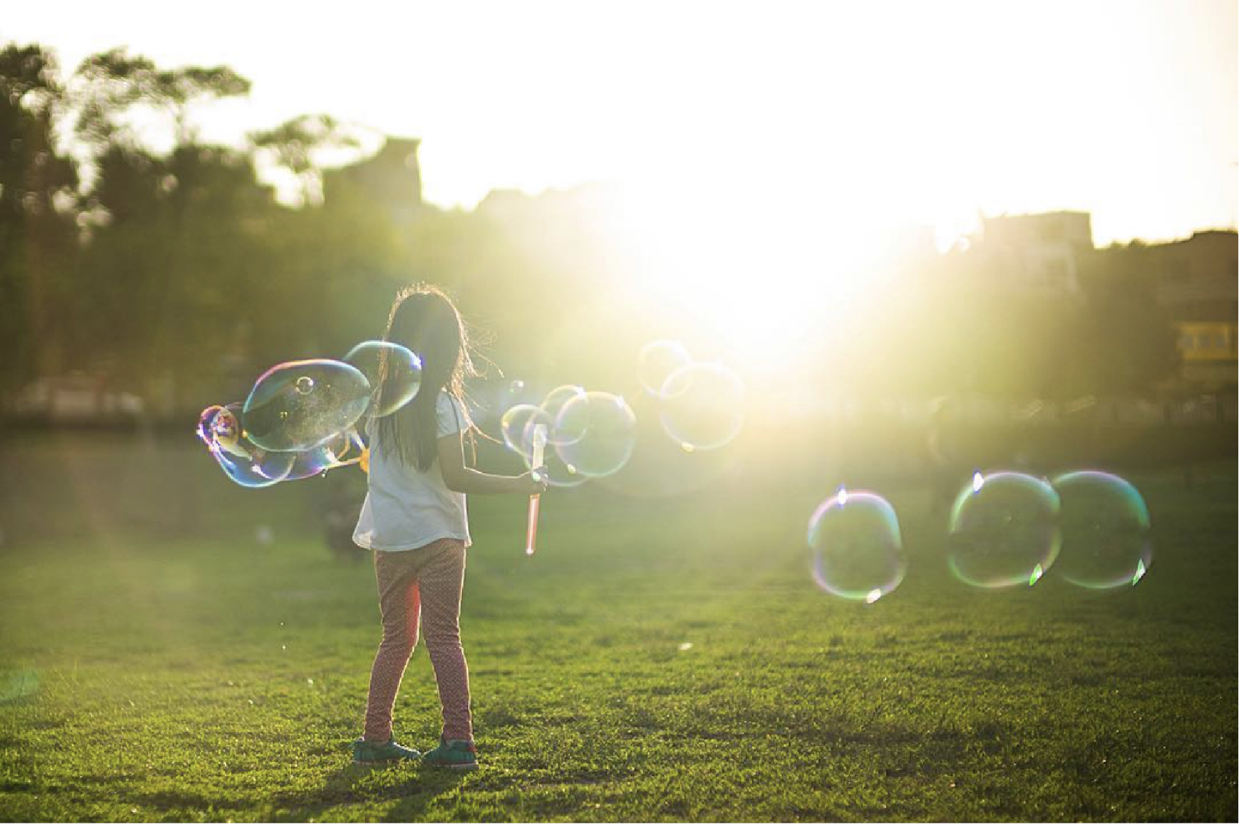 A girl is standing in a grassy field. The background is blurred, but shows some trees and buildings, and a bright sun, just behind the buidlings. The girl is facing toward the buildings, and appears to be turning. Her toes are pointed toward each other and she is holding a wand from which a dozen or so large head-sized bubbles have come, floating around her.