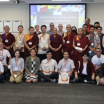 A large group of people sitting and standing in two rows, a large screen behind them
