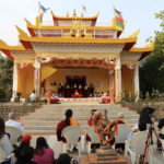 The Buddha Hall temple at Deer Park in Bir, India is shown with some performers on a stage under the awning. Rinpoche is seated in the center of a couple of rows of audience members, just behind the statue of Saraswati, holding his phone as if taking a photo or video of the performance.