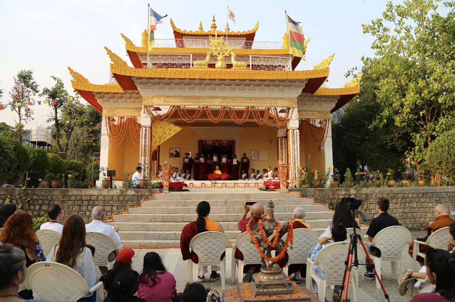 The Buddha Hall temple at Deer Park in Bir, India is shown with some performers on a stage under the awning. Rinpoche is seated in the center of a couple of rows of audience members, just behind the statue of Saraswati, holding his phone as if taking a photo or video of the performance.